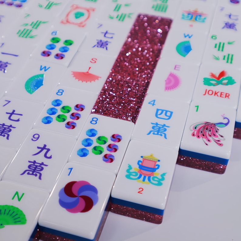 May include: A collection of white Mahjong tiles featuring colorful printed designs including Chinese characters, numbers, fans, a peacock, and a joker mask. The tiles have a dark blue base and a strip of pink glitter embedded along the side.