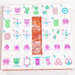 Puede incluir: Un conjunto de fichas de mahjong blancas dispuestas en una cuadr&iacute;cula, con dise&ntilde;os impresos coloridos que incluyen flores de loto, tallos de bamb&uacute;, m&aacute;scaras de comod&iacute;n, n&uacute;meros y caracteres chinos. Tres fichas en la columna central tienen un acabado naranja marmoleado.