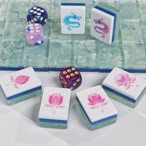 May include: A set of mahjong tiles with translucent light green bases and white tops featuring pink lotus flowers numbered one through four, blue dragon illustrations, and three multi-coloured dice with white or gold pips.