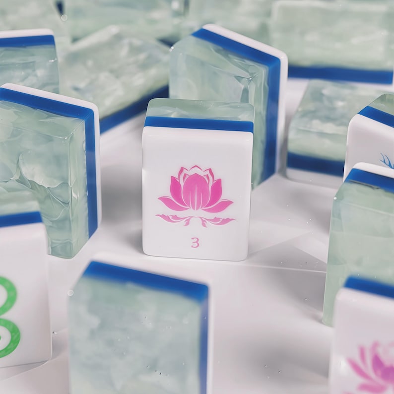 May include: A collection of rectangular mahjong tiles featuring a white front face with a pink lotus flower graphic and the number 3. The tiles have a translucent, pale green, marbled back section separated from the white front by a blue stripe.