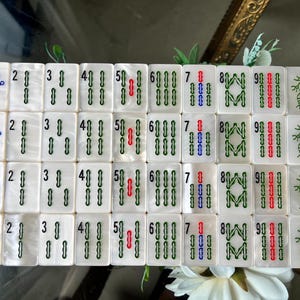 May include: A set of white rectangular mahjong tiles arranged in a grid, featuring engraved numbers one through nine in green, blue, and red, alongside bird and dragon illustrations, all with a pearlescent finish on the surface of each tile.
