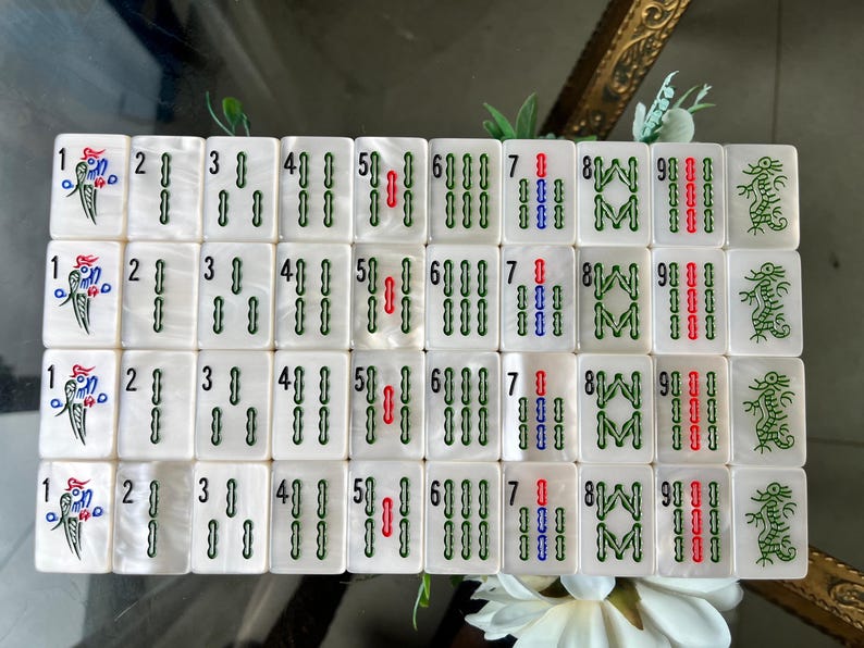 May include: A set of white rectangular mahjong tiles arranged in a grid, featuring engraved numbers one through nine in green, blue, and red, alongside bird and dragon illustrations, all with a pearlescent finish on the surface of each tile.
