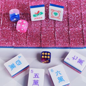 May include: A set of Mahjong tiles with pink glitter backs and blue bases, featuring white faces with various symbols, numbers, and a Joker tile. Two dice are included, one pink with white dots and one blue and purple with gold dots.