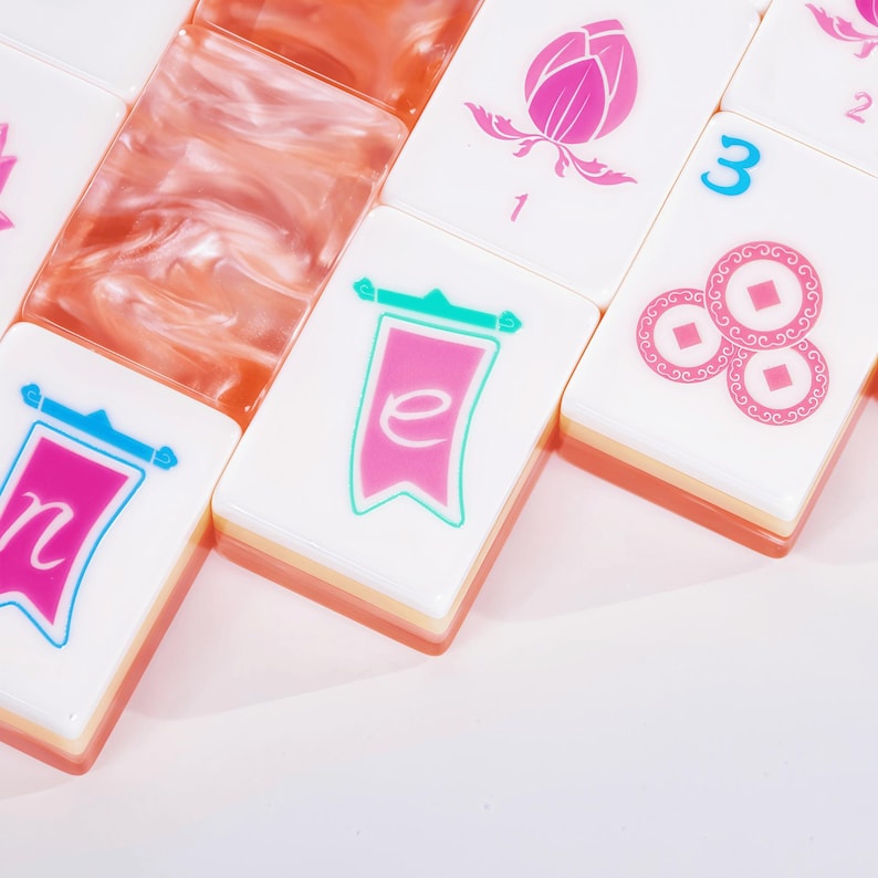Puede incluir: Un conjunto de fichas de mahjong rectangulares con caras blancas y dise&ntilde;os impresos en rosa y azul, que incluyen estandartes con letras, una flor de loto con el n&uacute;mero 1 y tres monedas con el n&uacute;mero 3, junto a fichas de color melocot&oacute;n transl&uacute;cido.