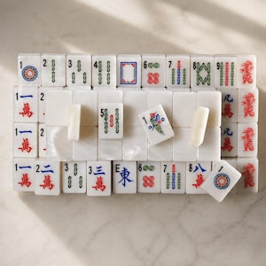 May include: A collection of white rectangular Mahjong tiles arranged in rows. The tiles feature engraved symbols, numbers, and characters in red, green, and blue ink. Some tiles are placed horizontally, while others are upright, displaying various suits.