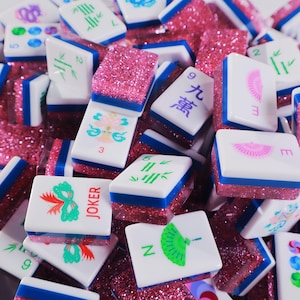 May include: A collection of Mahjong tiles featuring white faces with colorful symbols, numbers, and the word JOKER, set against a base of sparkling pink glitter. Each tile has a blue horizontal stripe separating the white top from the pink base.