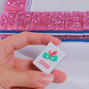 May include: A white rectangular Mahjong tile featuring a green mask graphic and the red text JOKER, held in front of a row of pink glittery tiles with blue bases. The tile has a smooth finish and clear, bold lettering for easy identification.