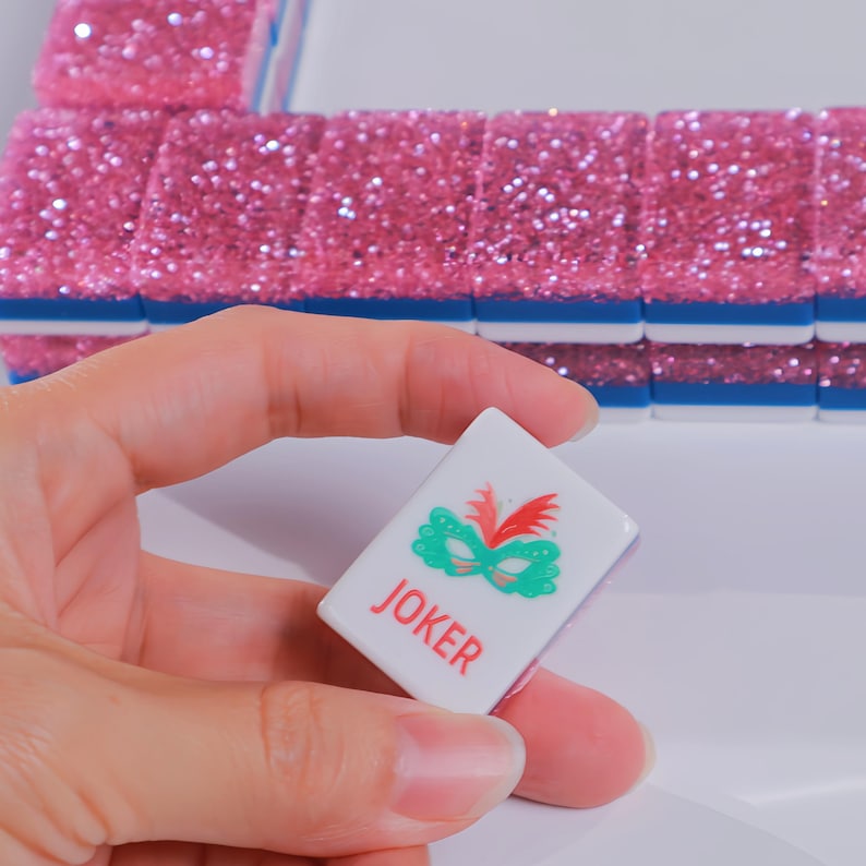 May include: A white rectangular Mahjong tile featuring a green mask graphic and the red text JOKER, held in front of a row of pink glittery tiles with blue bases. The tile has a smooth finish and clear, bold lettering for easy identification.