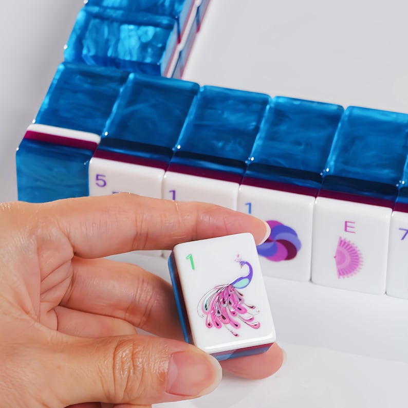 May include: A set of rectangular Mahjong tiles with translucent blue marbled backs and white fronts. The tiles feature various printed designs including a peacock with the number 1, a purple swirl, a pink fan, and the letter E, alongside numerical digits.