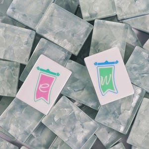 May include: A collection of rectangular mahjong tiles with a translucent, marbled light blue and white finish. Two tiles are placed on top, one featuring a pink banner graphic with a lowercase e, and the other a green banner with a lowercase w.