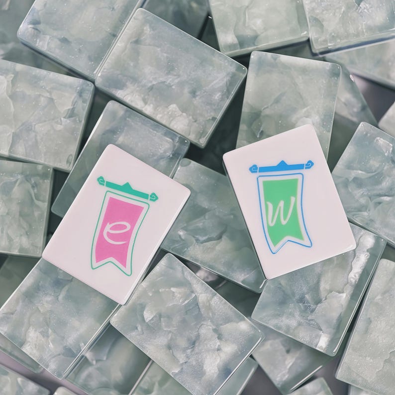 May include: A collection of rectangular mahjong tiles with a translucent, marbled light blue and white finish. Two tiles are placed on top, one featuring a pink banner graphic with a lowercase e, and the other a green banner with a lowercase w.