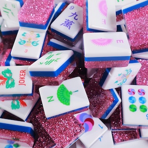 May include: A collection of Mahjong tiles featuring white faces with colorful printed designs, including fans, masks, and Chinese characters. Each tile has a blue middle stripe and a thick, textured base filled with sparkling pink glitter.