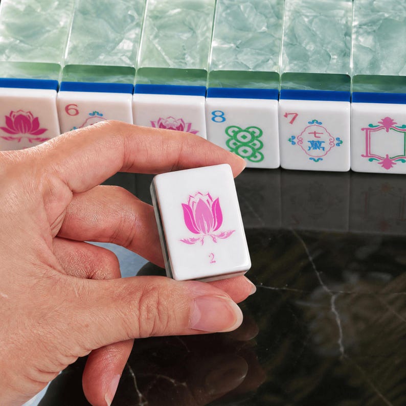 May include: A rectangular mahjong tile with a white face featuring a pink lotus flower graphic and the number 2 in pink. Behind it, a row of similar tiles with translucent green backs and various symbols, numbers, and floral designs in blue and pink.