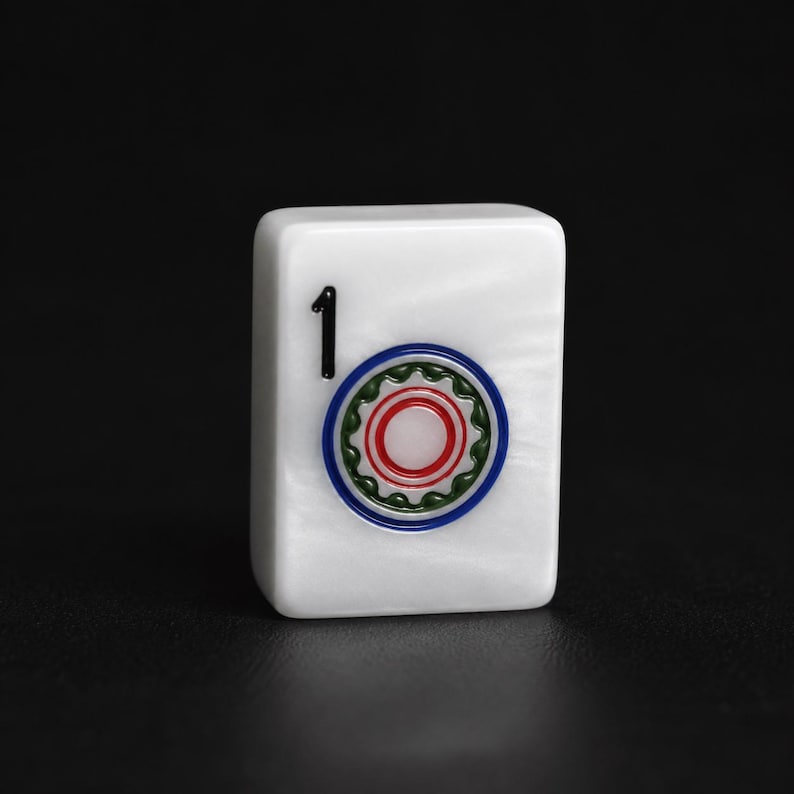 May include: A white rectangular mahjong tile with a pearlescent finish. The tile features the black number 1 in the upper left corner and a circular design in the center consisting of a red center, a green scalloped ring, and a blue outer border.