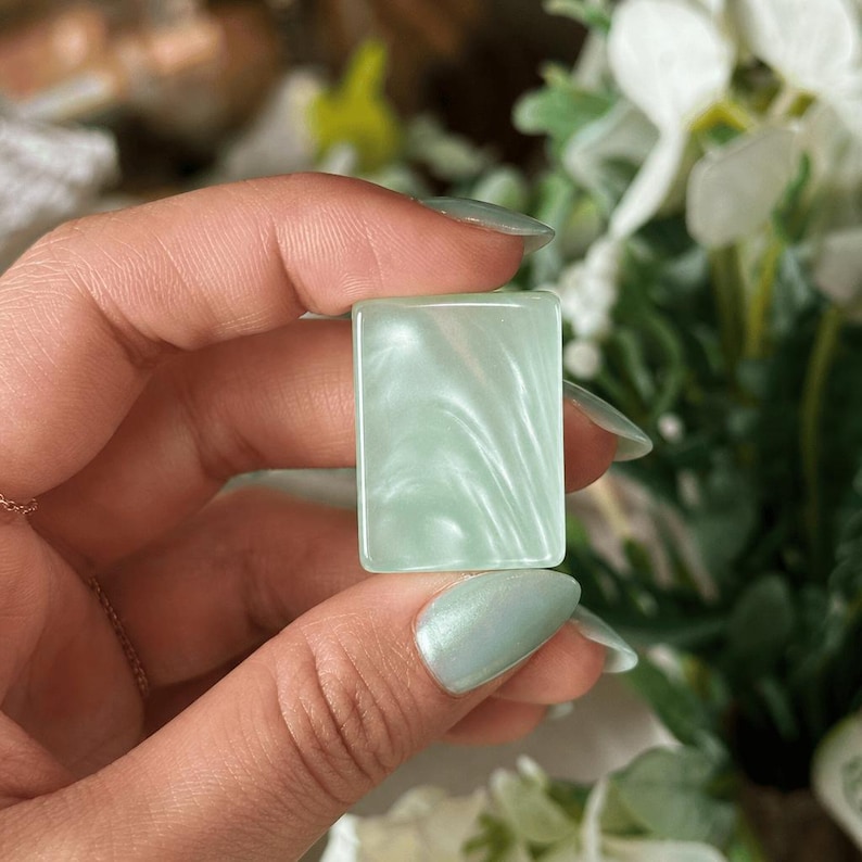 May include: A rectangular cabochon with a smooth, polished surface in a pale mint green color. The material features a swirling, pearlescent finish that creates subtle light reflections across the flat, slightly rounded edges of the piece.