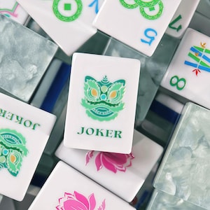 May include: A collection of white rectangular Mahjong tiles with various colourful designs. One central tile features a green lion head graphic above the word JOKER in green text. Other tiles display blue numbers, green bamboo patterns, and pink lotus flowers.