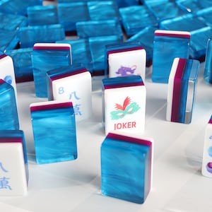 May include: A collection of rectangular mahjong tiles featuring a translucent blue marbled back and a white front face. Some tiles display colourful graphics, including a green mask with red feathers and the text JOKER, or the number 8 with Chinese characters.