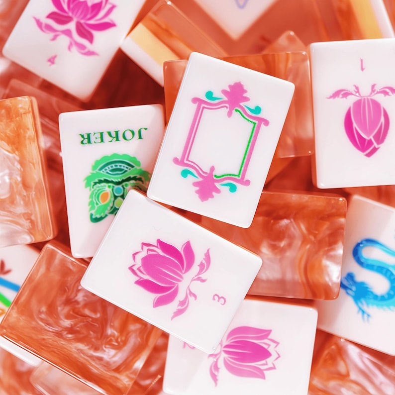 Puede incluir: Una colecci&oacute;n de fichas de mahjong rectangulares con caras blancas y dise&ntilde;os impresos coloridos, que incluyen flores de loto rosas, una cabeza de drag&oacute;n verde y naranja, un drag&oacute;n azul y un marco rosa. El reverso de las fichas es de resina naranja veteada.
