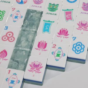 May include: A collection of white mahjong tiles featuring colourful printed designs including pink lotus flowers, blue and red Chinese characters, green lion heads, and the word JOKER. Some tiles have blue bases, and a row of translucent light blue tiles.