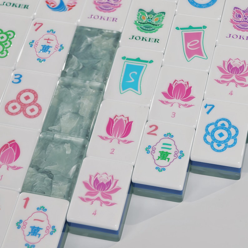 May include: A collection of white mahjong tiles featuring colourful printed designs including pink lotus flowers, blue and red Chinese characters, green lion heads, and the word JOKER. Some tiles have blue bases, and a row of translucent light blue tiles.