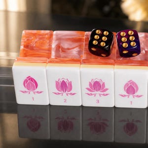 Puede incluir: Un conjunto de fichas de mahjong rectangulares con caras blancas que presentan ilustraciones de flores de loto rosas y los n&uacute;meros 1, 2 y 3. Las fichas tienen dorsos jaspeados de color naranja y est&aacute;n coronadas por dos dados transl&uacute;cidos, uno negro y otro morado, con puntos dorados.