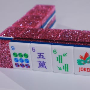 May include: A set of Mahjong tiles with a vibrant pink glitter finish on the back. The front faces are white with blue edges, displaying various symbols including the number 9, the Chinese character for five, the number 6, and a green mask with JOKER.