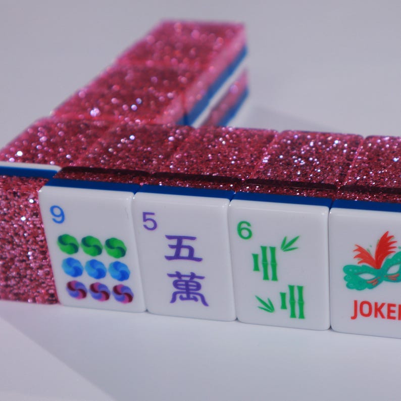 May include: A set of Mahjong tiles with a vibrant pink glitter finish on the back. The front faces are white with blue edges, displaying various symbols including the number 9, the Chinese character for five, the number 6, and a green mask with JOKER.