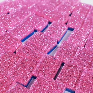 May include: A collection of rectangular dominoes featuring a vibrant pink glitter finish on the top surface. Each piece has a solid blue base layer visible along the sides, creating a distinct contrast between the sparkling pink top and the blue bottom.