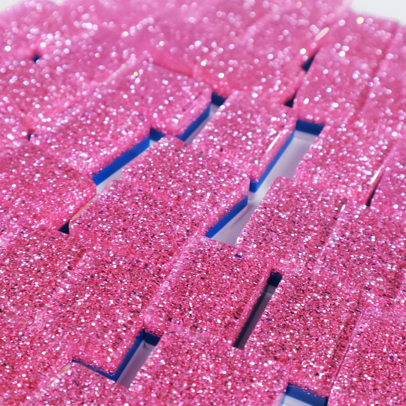 May include: A collection of rectangular dominoes featuring a vibrant pink glitter finish on the top surface. Each piece has a solid blue base layer visible along the sides, creating a distinct contrast between the sparkling pink top and the blue bottom.