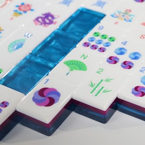 May include: A set of white Mahjong tiles with colourful graphic designs including fans, bamboo, numbers, and swirling patterns. The tiles feature a layered construction with a translucent blue base and a dark purple middle section, arranged in a grid.