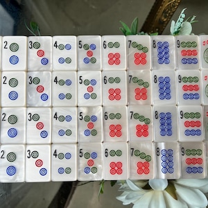 May include: A set of white rectangular mahjong tiles arranged in four rows. Each tile features a number from one to nine or a joker symbol. The designs consist of colorful concentric circles in red, blue, and green, with the word JOKER in red ink.