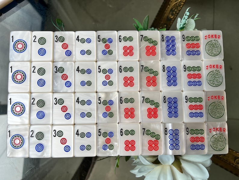 May include: A set of white rectangular mahjong tiles arranged in four rows. Each tile features a number from one to nine or a joker symbol. The designs consist of colorful concentric circles in red, blue, and green, with the word JOKER in red ink.
