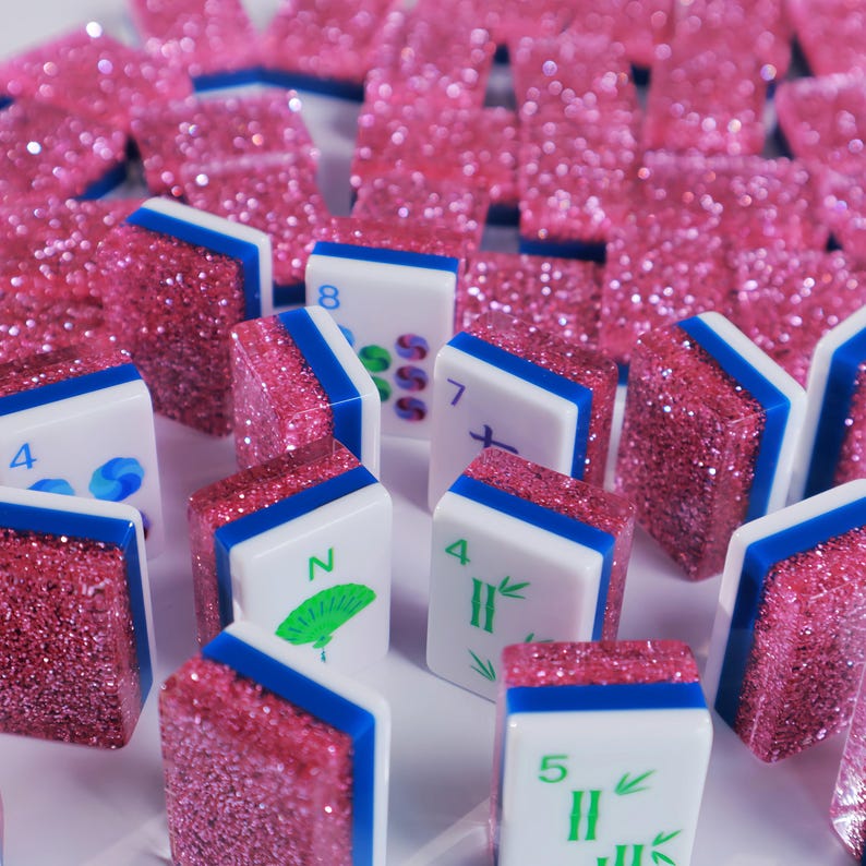 May include: A collection of Mahjong tiles featuring white faces with green and blue printed numbers and symbols, accented by a thin blue stripe and a thick, textured, sparkling pink glitter backing on each individual rectangular game piece.