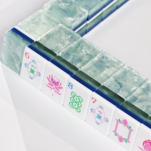 May include: A set of mahjong tiles featuring a translucent, marbled light blue and white crystalline texture on the back. The front faces are white with colourful engraved symbols, including a pink lotus, green circles, and Chinese characters.