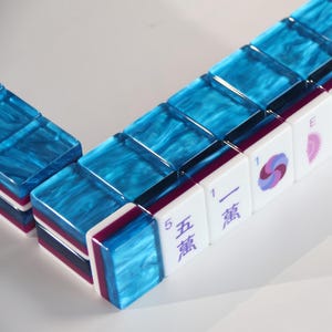 May include: A set of rectangular mahjong tiles arranged in a row. Each tile features a vibrant blue marbled top surface, a dark purple middle layer, and a white base. Some tiles display purple Chinese characters, numbers, and circular floral symbols.
