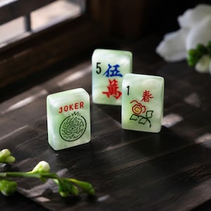 May include: Three rectangular mahjong tiles made of a pale green, marbled material. One tile features the word JOKER in red with a green circular graphic, another shows the number 5 and blue Chinese characters, and the third displays the number 1.