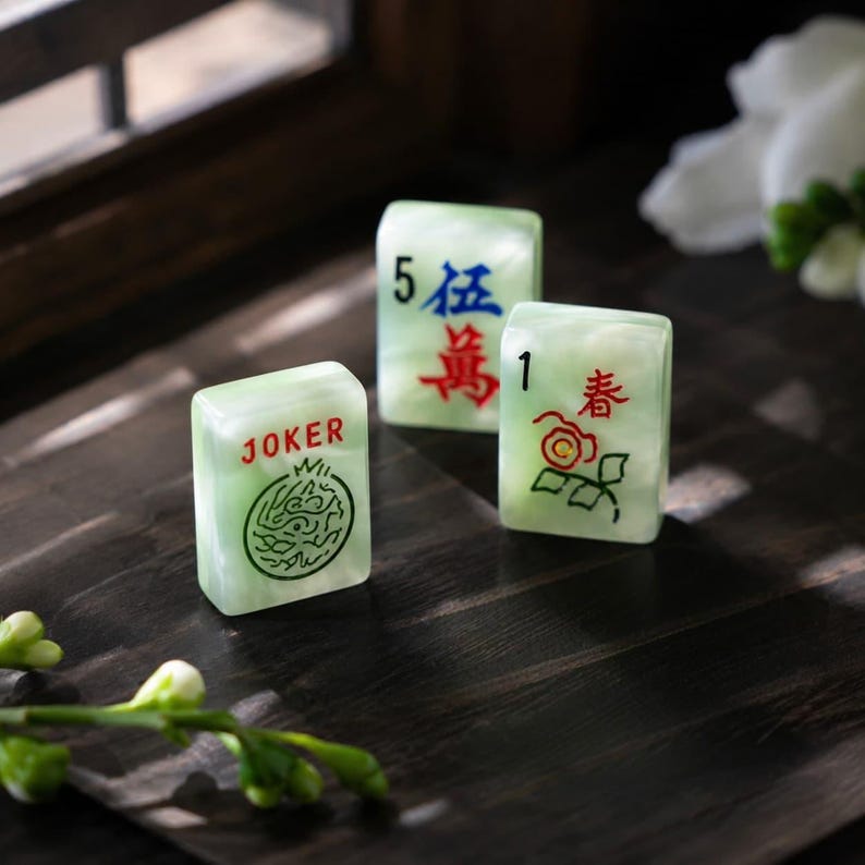 May include: Three rectangular mahjong tiles made of a pale green, marbled material. One tile features the word JOKER in red with a green circular graphic, another shows the number 5 and blue Chinese characters, and the third displays the number 1.