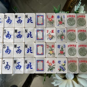 May include: A set of rectangular Mahjong tiles with a pearlescent white finish. The tiles feature blue engraved Chinese characters for compass directions, red and blue geometric frames, floral illustrations with numbers, and green Joker tiles.