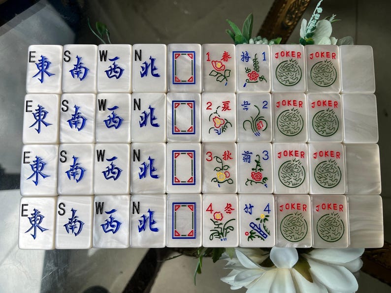 May include: A set of rectangular Mahjong tiles with a pearlescent white finish. The tiles feature blue engraved Chinese characters for compass directions, red and blue geometric frames, floral illustrations with numbers, and green Joker tiles.