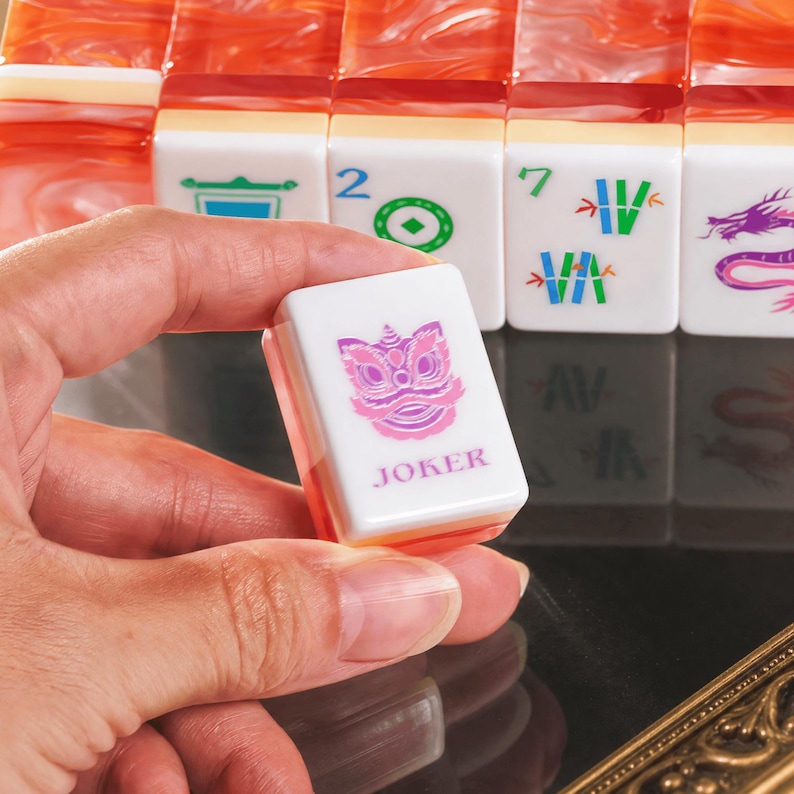 Puede incluir: Una ficha de Mahjong blanca con un gr&aacute;fico de le&oacute;n danzante morado y la palabra JOKER en texto morado, sostenida frente a una fila de fichas de Mahjong de color naranja jaspeado con varios s&iacute;mbolos y n&uacute;meros verdes, azules y morados.