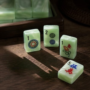 May include: Four pale green rectangular mahjong tiles with smooth surfaces. One tile shows a red and green circular pattern with the number 1, another shows two blue circles with the number 2, one features a red flower with the number 1, and one shows red and blue Chinese characters with the number 5.