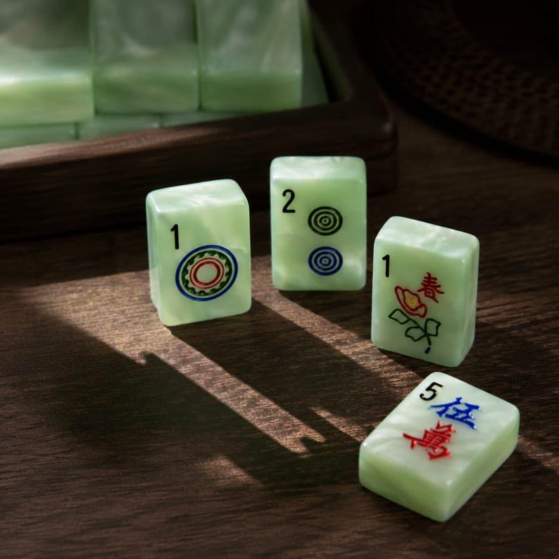 May include: Four pale green rectangular mahjong tiles with smooth surfaces. One tile shows a red and green circular pattern with the number 1, another shows two blue circles with the number 2, one features a red flower with the number 1, and one shows red and blue Chinese characters with the number 5.