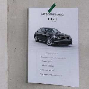 May include: A white paper with the text "MERCEDES-AMG C63 (W204)" and car specifications, including engine size, horsepower, torque, and acceleration. The image shows a black sedan. The paper is attached to a concrete wall.