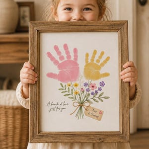 Op de afbeelding: Een rechthoekige houten lijst met een afdruk van twee handafdrukken, één roze en één geel, gerangschikt als een bloemenboeket met groene stelen en kleine kleurrijke bloemen. De tekst luidt A bunch of love just for you en Ava & Daniel.
