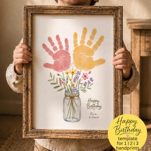May include: A framed art print featuring a handprint craft design with a pink handprint and a yellow handprint above a mason jar filled with colourful wildflowers. The text reads Happy Birthday, Olivia & Daniel, with a yellow circular sticker.