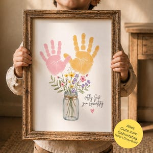 Op de afbeelding: Een ingelijste print met roze en gele handafdrukken, een bloemstuk in een pot en de tekst "Alles Gute zum Geburtstag". De print wordt vastgehouden door een kind.