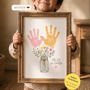 Op de afbeelding: Een ingelijste print met roze en gele handafdrukken, een bloemstuk in een glazen pot en Duitse tekst. Het kind houdt de print vast. De lijst is bruin.
