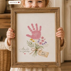 May include: A framed print with a pink handprint and a floral bouquet. The print has script text and a tag that reads "Deine Sophie". The wooden frame adds a rustic touch to the artwork.