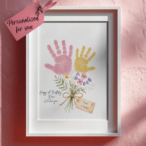 Könnte beinhalten: Ein gerahmter Druck mit rosa und gelben Handabdrücken, die einen Blumenstrauß bilden, mit zusätzlichen Blumen. Der Druck enthält den Text "Happy 1st Birthday, Nana, We love you" und ein Etikett mit der Aufschrift "Daniel & Sophia". Ein rosa Etikett lautet "Personalisiert für dich".