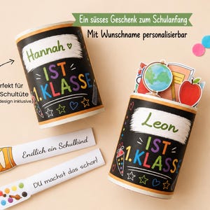 May include: Two cylindrical black containers with chalkboard-style graphics, featuring the text 'Hannah IST 1. KLASSE' and 'Leon IST 1. KLASSE'. Includes two white rectangular tags with illustrations of crayons and paint, and text 'Endlich ein Schulkind' and 'DU machst das schon'.